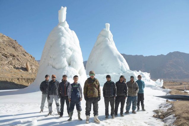 A group of students, dressed in warm coats and winter pants, stand in front of two large domes of ice.