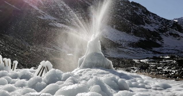 A dome-shaped block of ice, surrounded by snow and ice, sprays water into the air from its top. 